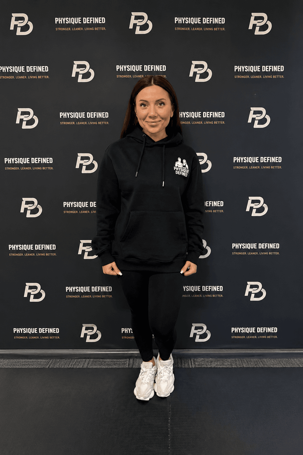 Woman in black hoodie and leggings posing against a repeating Physique Defined logo backdrop.