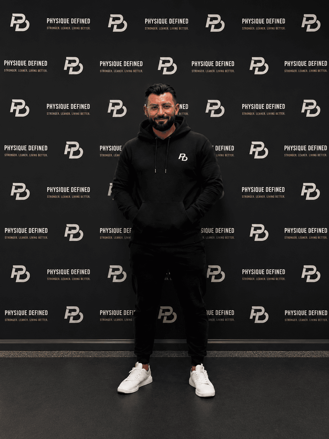 Bearded man in black athletic wear posing against a black Physique Defined branded backdrop.